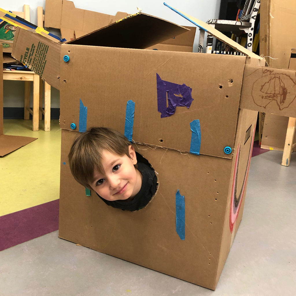 Child in a cardboard box with his head sticking out of a hole in the side.