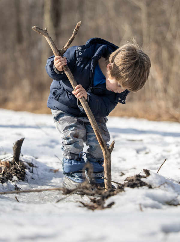 Child poking large stick into snow covered leaves.