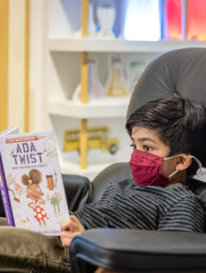 You child sits in a chair reading the book 'Ada Twist, Scientist' written by Andrea Beaty and illustrated by David Roberts.