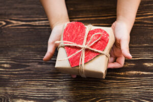 Child's hands holding a small package wrapped in brown paper, with a colored-in red heart, secured with a twine bow.